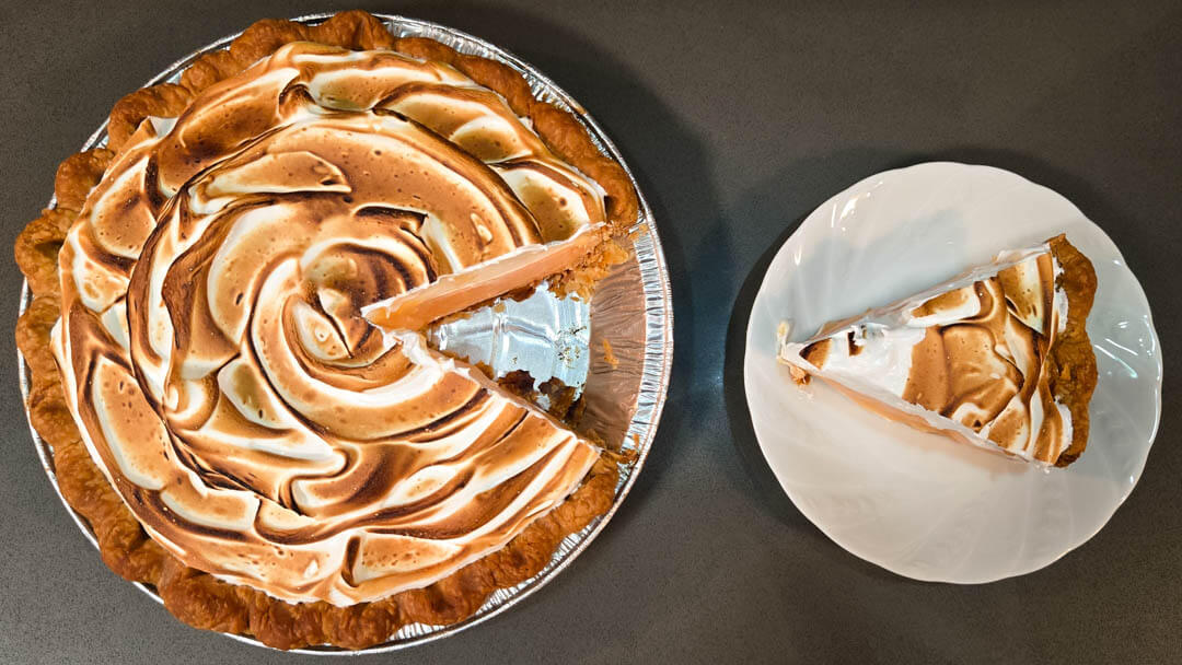 overhead photo of a grapefruit meringue pie with pale pink grapefruit curd in a flaky pie crust with toasted meringue topping. A slice has been cut out and placed on a white plate next to it.