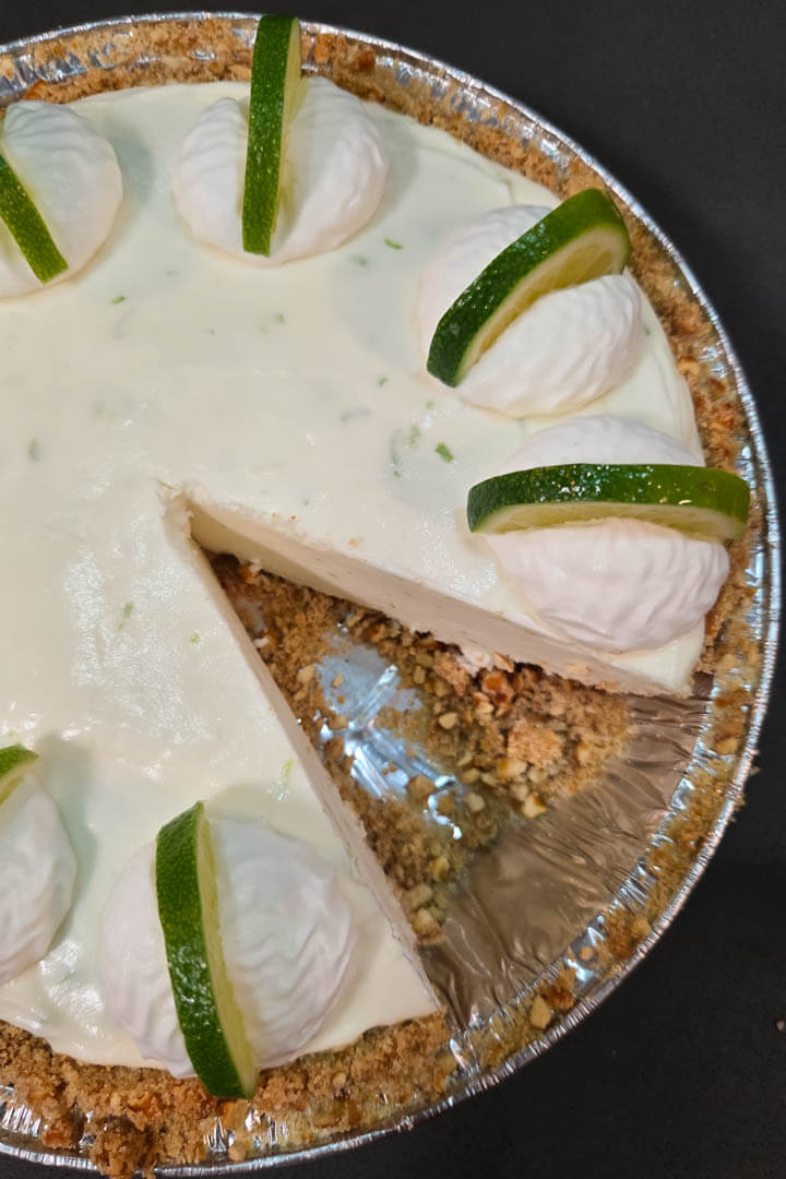overhead photo of a frozen margarita pie with a slice cut out. Cream margarita ice cream in a crunchy pie crust made with crushed pretzels decorated with whipped cream and fresh lime slices