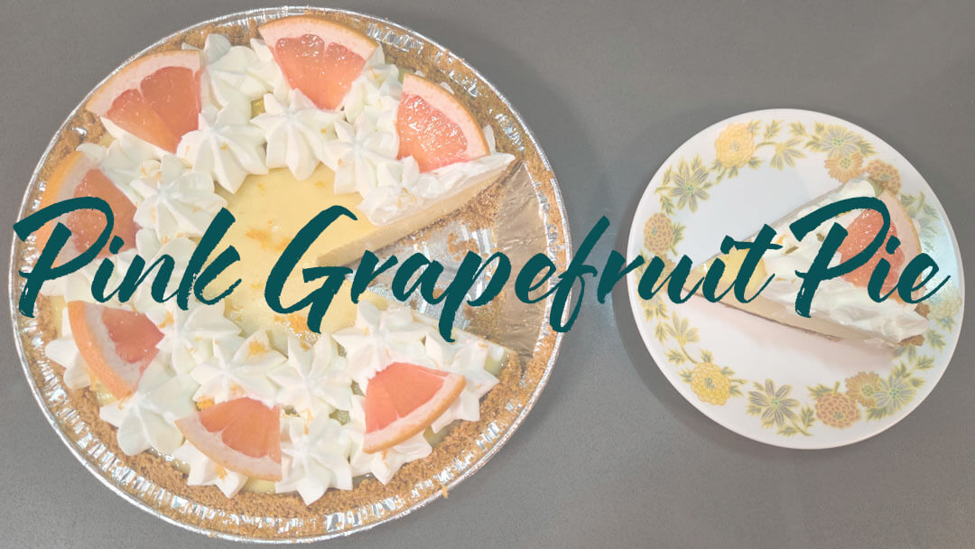 Pink grapefruit pie with grapefruit pie filling in a graham cracker pie crust decorated with grapefruit zest, grapefruit slices, and piped whipped cream. A slice is cut out and placed next to the pie on a white plate with yellow flowers. Text overlay reads "Pink Grapefruit Pie"