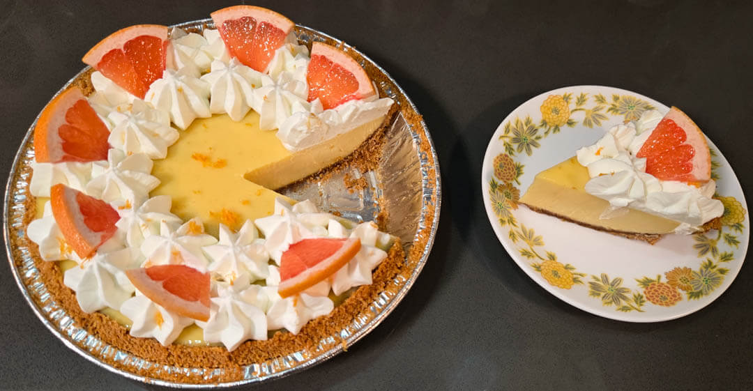 Pink grapefruit pie with grapefruit pie filling in a graham cracker pie crust decorated with grapefruit zest, grapefruit slices, and piped whipped cream. A slice is cut out and placed next to the pie on a white plate with yellow flowers