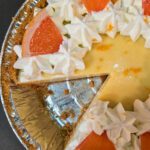 Pink grapefruit pie overhead photo in an aluminum pie tin. The icebox pie is decorated with whorls of fresh whipped cream, grapefruit zest, and slices of grapefruit. Text overlay reads "Pink Grapefruit Pie, As Easy as Key Lime Pie!" with the Doggone Baking logo in the corner