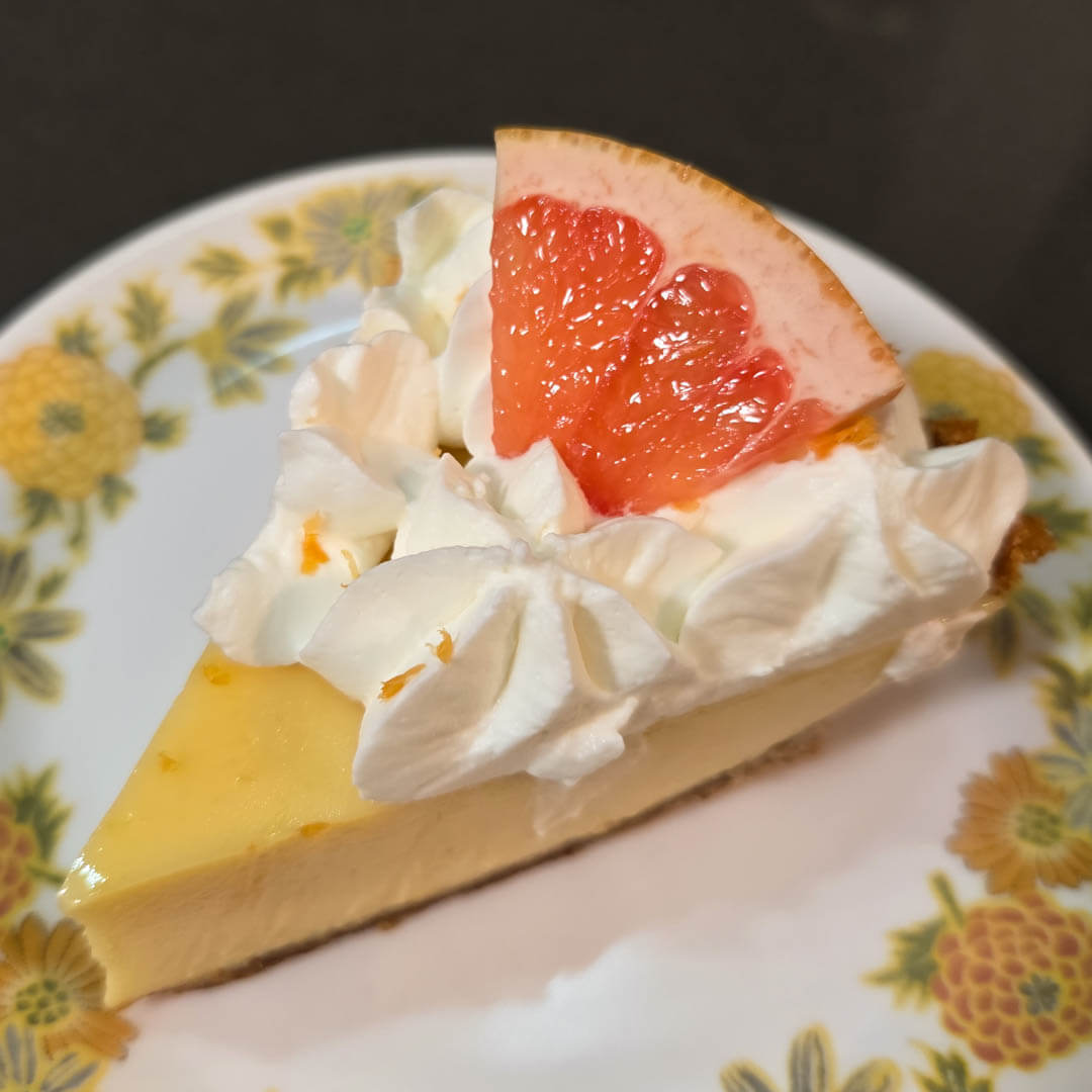 A slice of pink grapefruit pie on a white plate with yellow flowers. Graham cracker crust and decorated with a slice of pink grapefruit and swirls of whipped cream