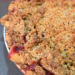 Overhead close-up of a strawberry rhubarb pie with a golden crumb topping and deep red fruit filling bubbling up through the crumble and around the crimped crust edges. Text overlay reads Strawberry Rhubarb Pie. Doggone Baking logo in corner.