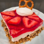 A Pinterest pin for a strawberry pretzel salad recipe. The image shows a close-up of a square slice of strawberry pretzel salad with a pretzel crumb crust, a cream cheese layer, and a strawberry jello topping with fresh strawberry slices, garnished with a dollop of whipped cream and a whole pretzel, with the Doggone Baking logo at the bottom. Text overlay reads "Strawberry Pretzel Salad | Potluck Dessert Recipe."