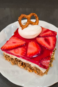 close up photo of a slice of strawberry pretzel salad with crunchy pretzel crust, sweet fluffy whipped cream filling, and strawberry jello topping with a dollop whipped cream and a pretzel on a white plate