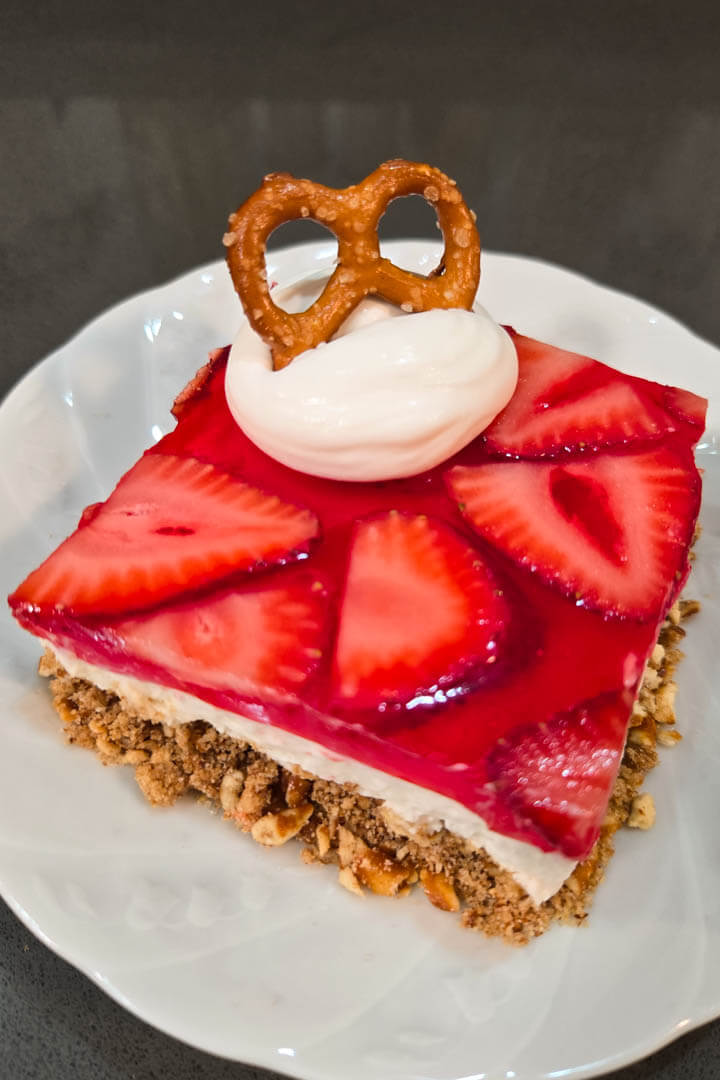 close up photo of a slice of strawberry pretzel salad with crunchy pretzel crust, sweet fluffy whipped cream filling, and strawberry jello topping with a dollop whipped cream and a pretzel on a white plate