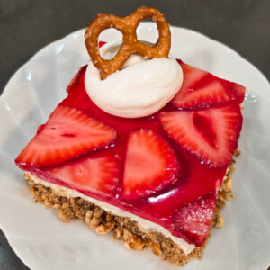 close up photo of a slice of strawberry pretzel salad with crunchy pretzel crust, sweet fluffy whipped cream filling, and strawberry jello topping with a dollop whipped cream and a pretzel on a white plate