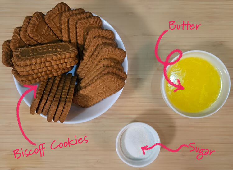 ingredients for biscoff crust laid out on a cutting board with text labels: biscoff cookies, sugar, butter