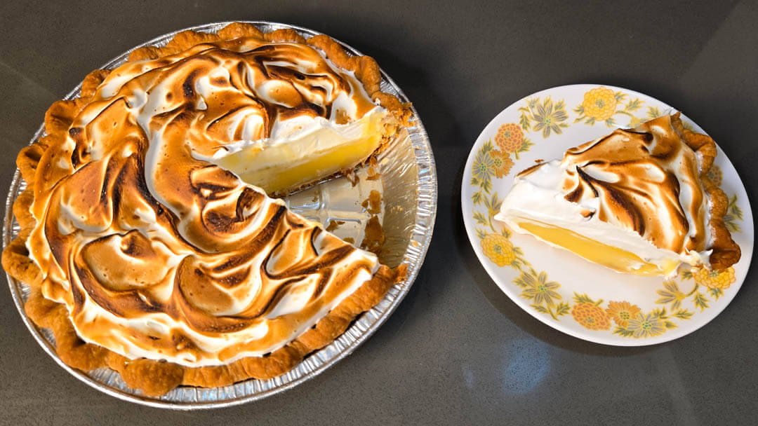 a photo of a fresh lemon meringue pie with a slice cut out in a metal pie tin with a golden crimped flaky pie crust, bright yellow lemon curd, and fluffy toasted meringue topping. The slice is placed on a plate next to the pie