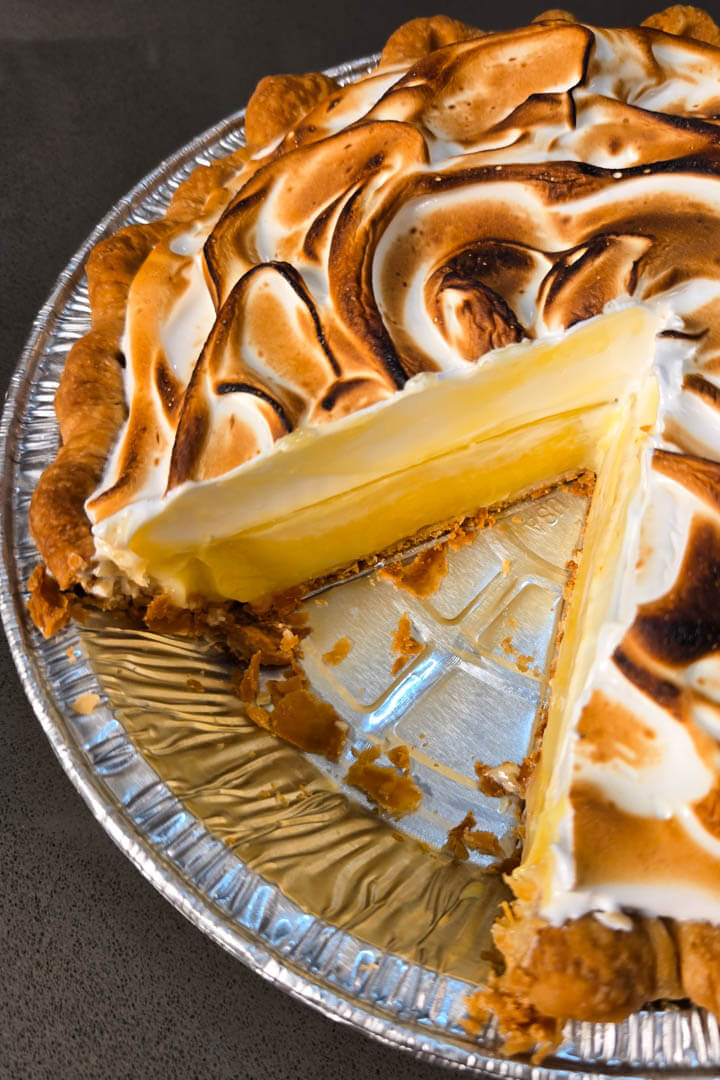 a photo of a classic lemon meringue pie with a slice cut out in a metal pie tin with a golden crimped flaky pie crust, bright yellow lemon curd, and fluffy toasted meringue topping