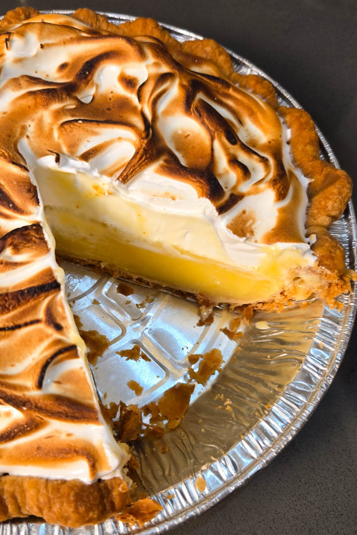 a photo of a fresh lemon meringue pie with a slice cut out in a metal pie tin with a golden crimped flaky pie crust, bright yellow lemon curd, and fluffy toasted meringue topping