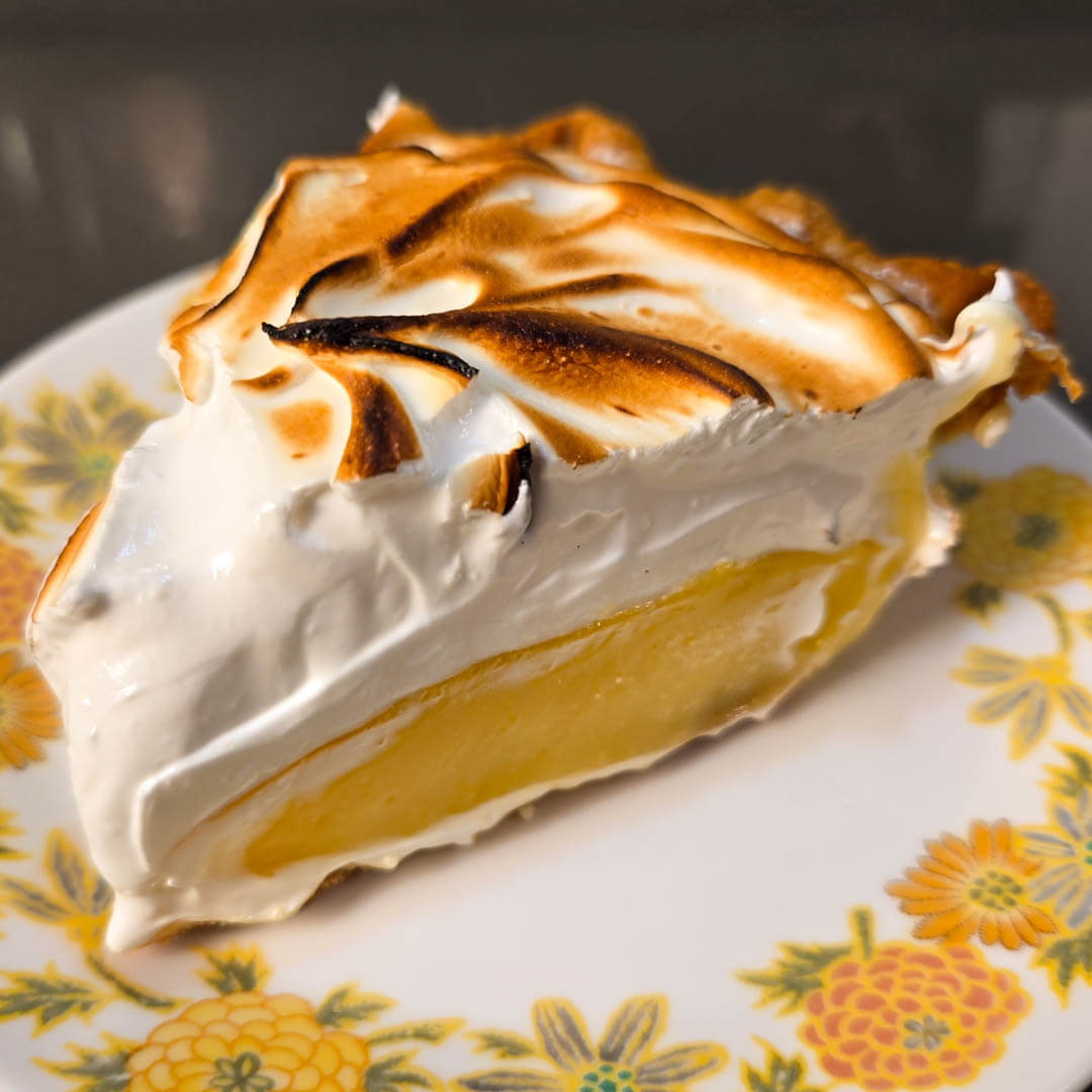 a slice of fresh lemon meringue pie with bright yellow lemon curd, fluffy toasted meringue topping, and a crimped pie crust on a floral plate