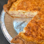 Overhead view of a homemade quiche Lorraine in a golden baked pastry shell with a slice removed, showing a thick, cheesy bacon and egg custard filling with a deeply golden, toasted top.