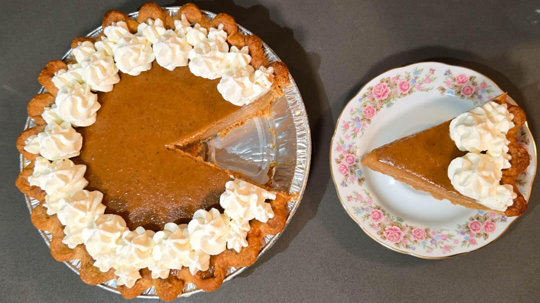 overhead photo of a whole pumpkin pie with whipped cream swirls and a slice cut out and placed next to the pie on a floral plate