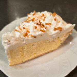 a slice of fluffy lilikoi chiffon pie with a pale yellow passionfruit mousse filling in a flaky pie crust topped with homemade whipped cream and toasted coconut