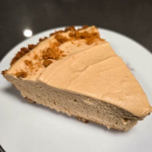 a slice of creamy cookie butter pie in a Biscoff pie crust decorated with cookie crumbles on a white plate with purple flowers