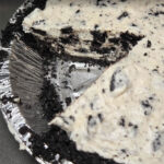 a photo of a no bake oreo pie with a slice cut out showing oreo pie crust and creamy no bake cookies and cream filling in an aluminum pie tin. Doggone Baking logo in left corner and text overlay reads "No bake dessert recipe! Cookies n' Cream pie"