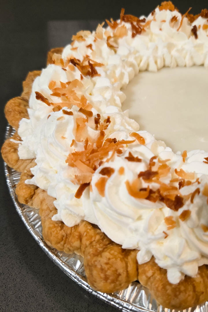 close up photo of a pumpkin haupia pie with crimped all-butter pie crust in an aluminum pie tin showing homemade whipped cream swirls topped with toasted shredded coconut