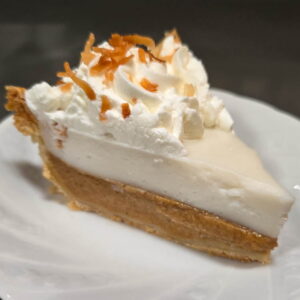 a slice of pumpkin haupia pie on a white plate showing creamy pumpkin filling in a flaky pie crust with a layer of haupia cream on top, topped with whipped cream swirls sprinkled with toasted coconut